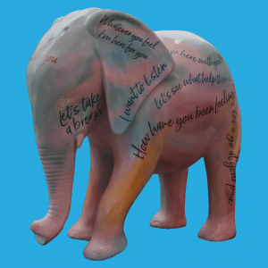 Elephant statue painted in shades of pink, blue, and green with mental health slogans written all over the statue. The background of the image is blue.