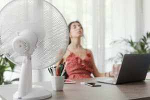 Woman sitting at desk at home and working with a computer, she is cooling herself with an electric fan